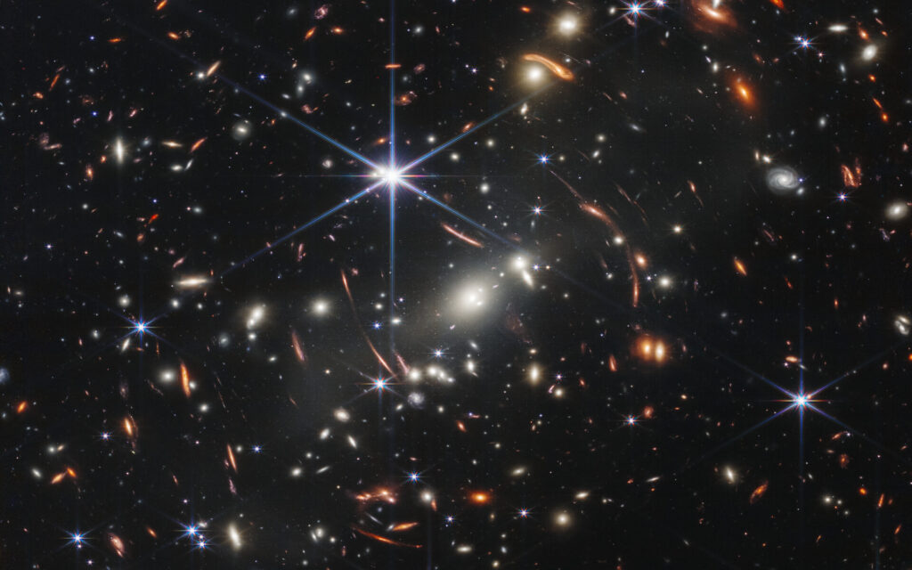 Webb First Deep Field of SMACS 0723: bright foreground stars, a galaxy cluster and orange arcs of gravitationally lensed background galaxies.